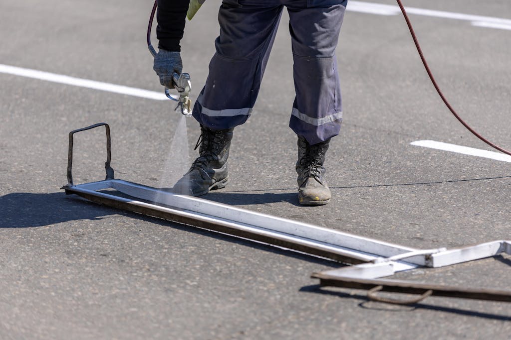 A construction worker painting white lines on a road with a spray gun during the day.