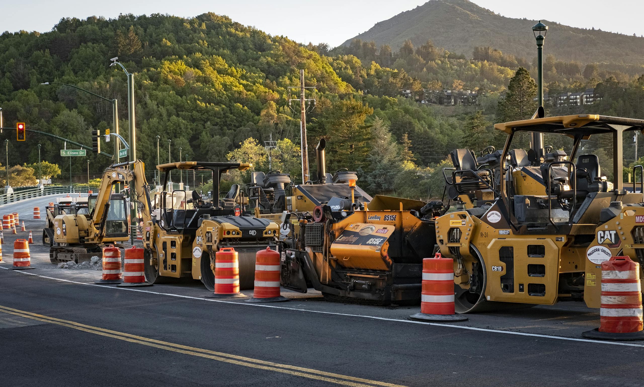 Road construction site with machinery and traffic cones under a scenic hill at sunset.